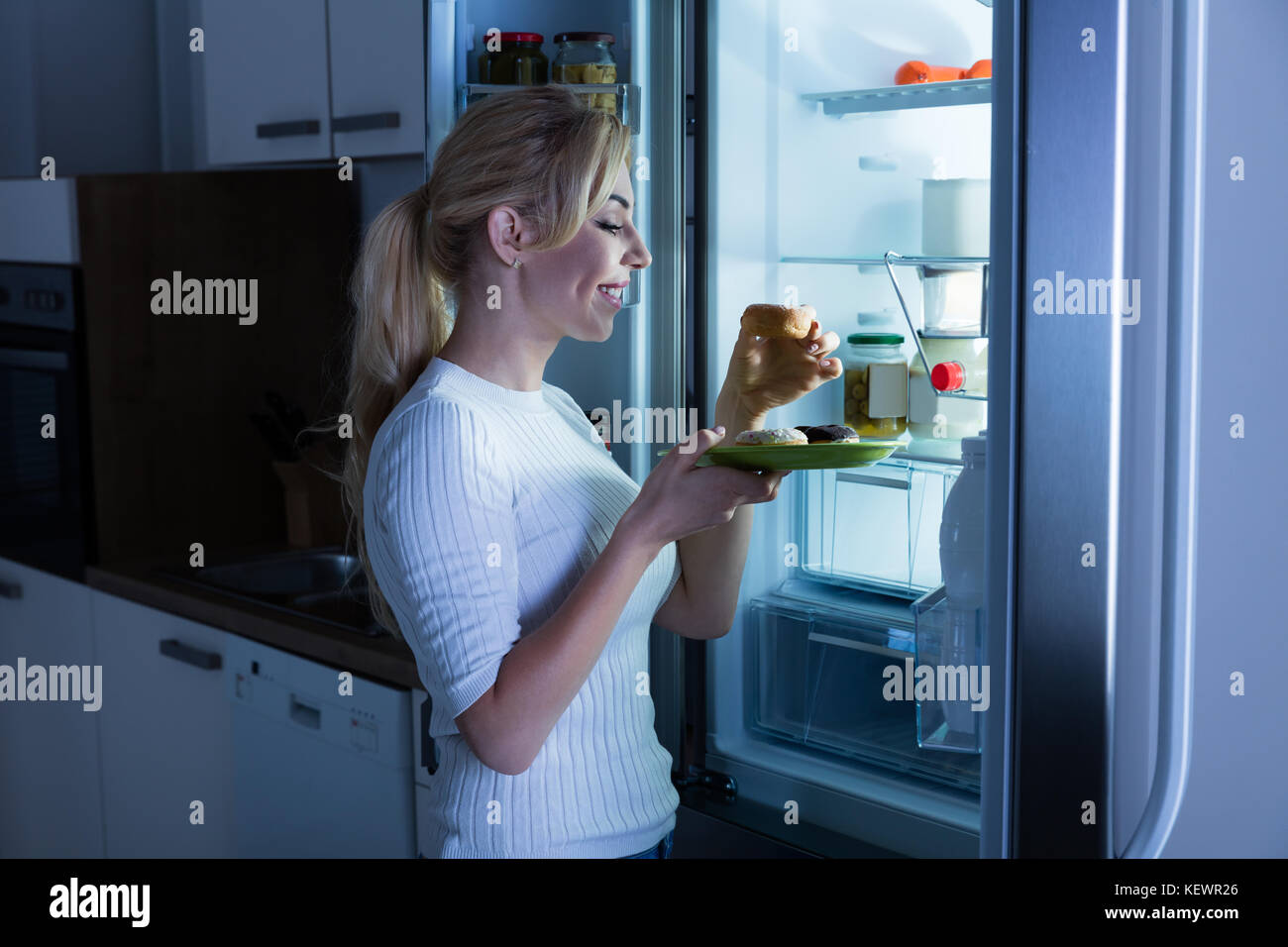 Jeune femme heureuse de manger un aliment sucré près de réfrigérateur dans la cuisine Banque D'Images