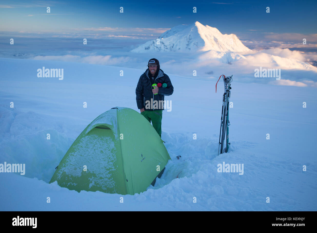 Un ski mountaineer jouit de la soirée vue sur le mont Hunter tout en vous tenant à côté de sa tente dans le camp de 14,000 pieds sur le glacier de Denali. Banque D'Images