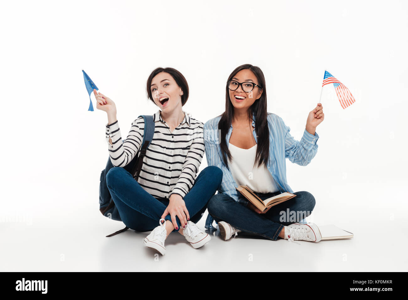 Deux jeunes étudiantes gaies holding drapeaux américains tout en restant assis ensemble sur le plancher isolé sur fond blanc Banque D'Images