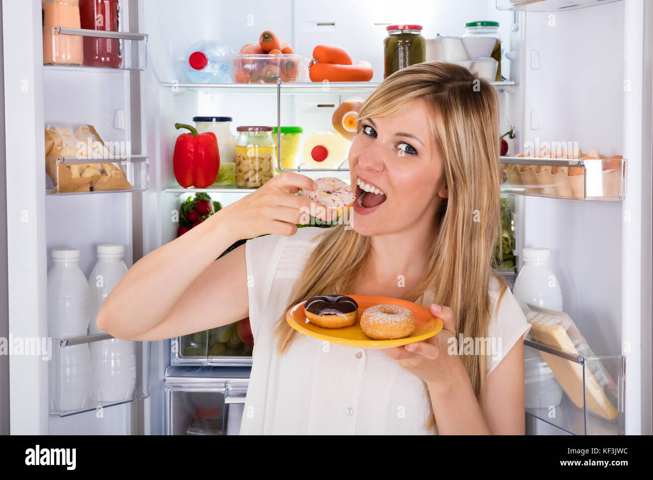 Young Happy Woman Eating Donut sucré a ouvert près de réfrigérateur Banque D'Images