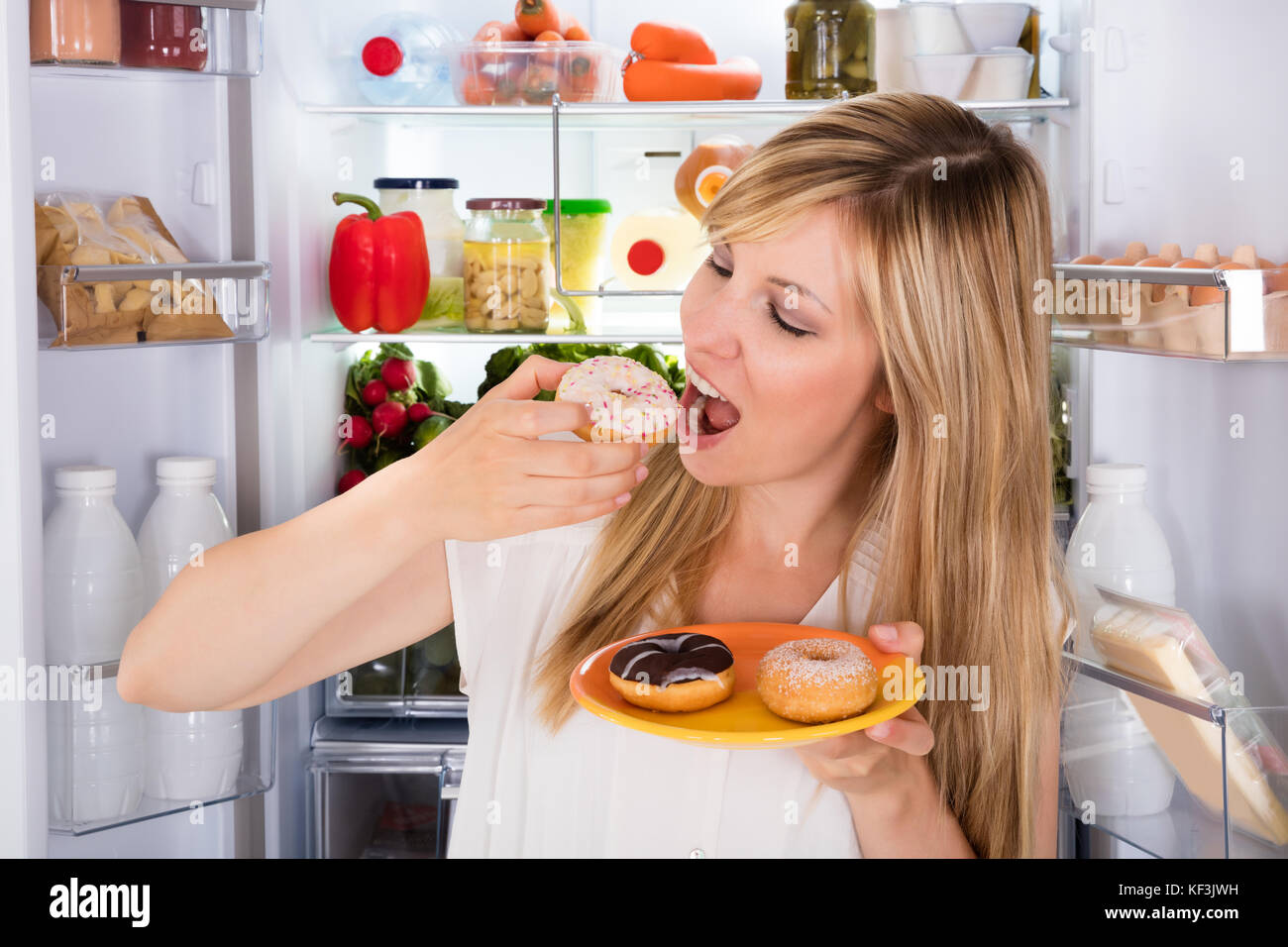 Young Happy Woman Eating Donut sucré a ouvert près de réfrigérateur Banque D'Images
