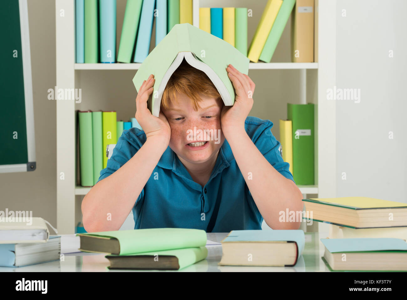 Angry Boy Sitting in Library Holding livre sur sa tête Banque D'Images