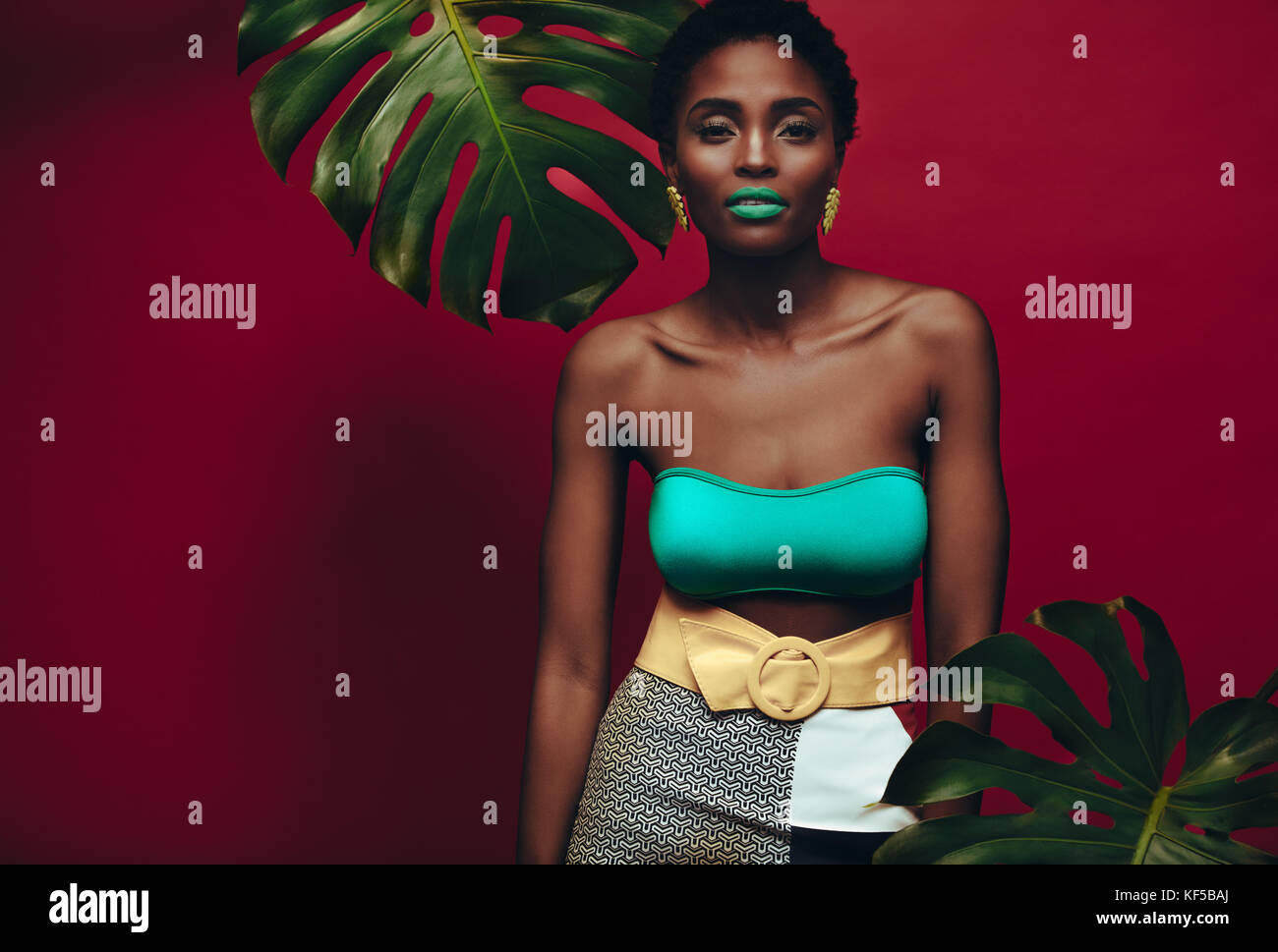 Portrait of female model aux couleurs maquillage sur fond rouge. Jeune femme africaine avec un miroir et des feuilles colorées. Banque D'Images