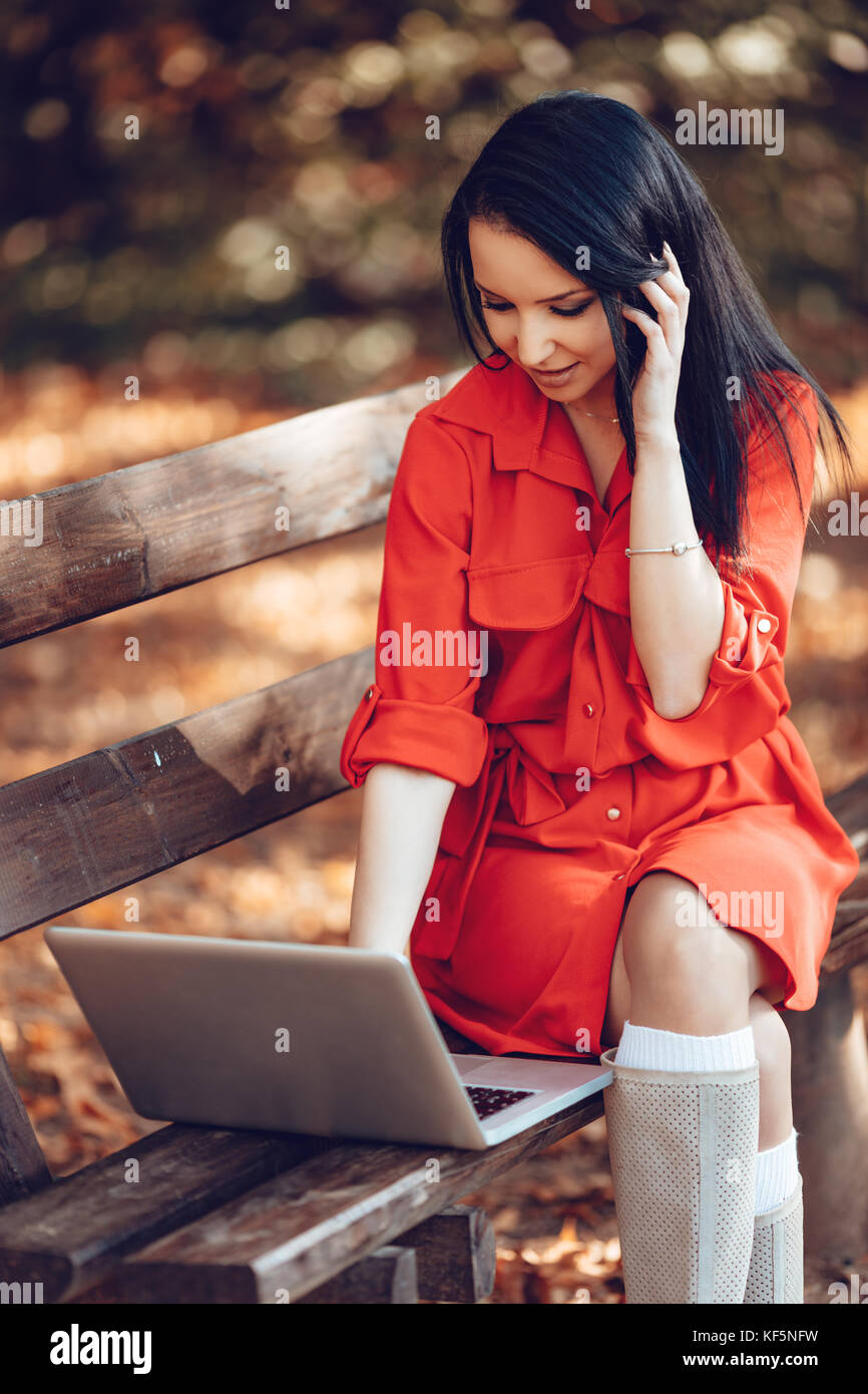 Jeune femme est à l'aide d'ordinateur portable dans le parc sur une journée ensoleillée d'automne, pendant une pause, et de parler sur son téléphone portable. Banque D'Images