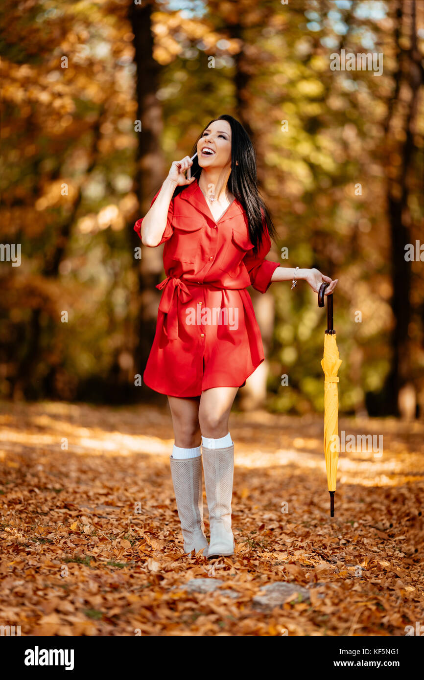 Jeune femme en robe rouge parler sur smartphone balade en automne sunny park, holding folded umbrella jaune. Banque D'Images