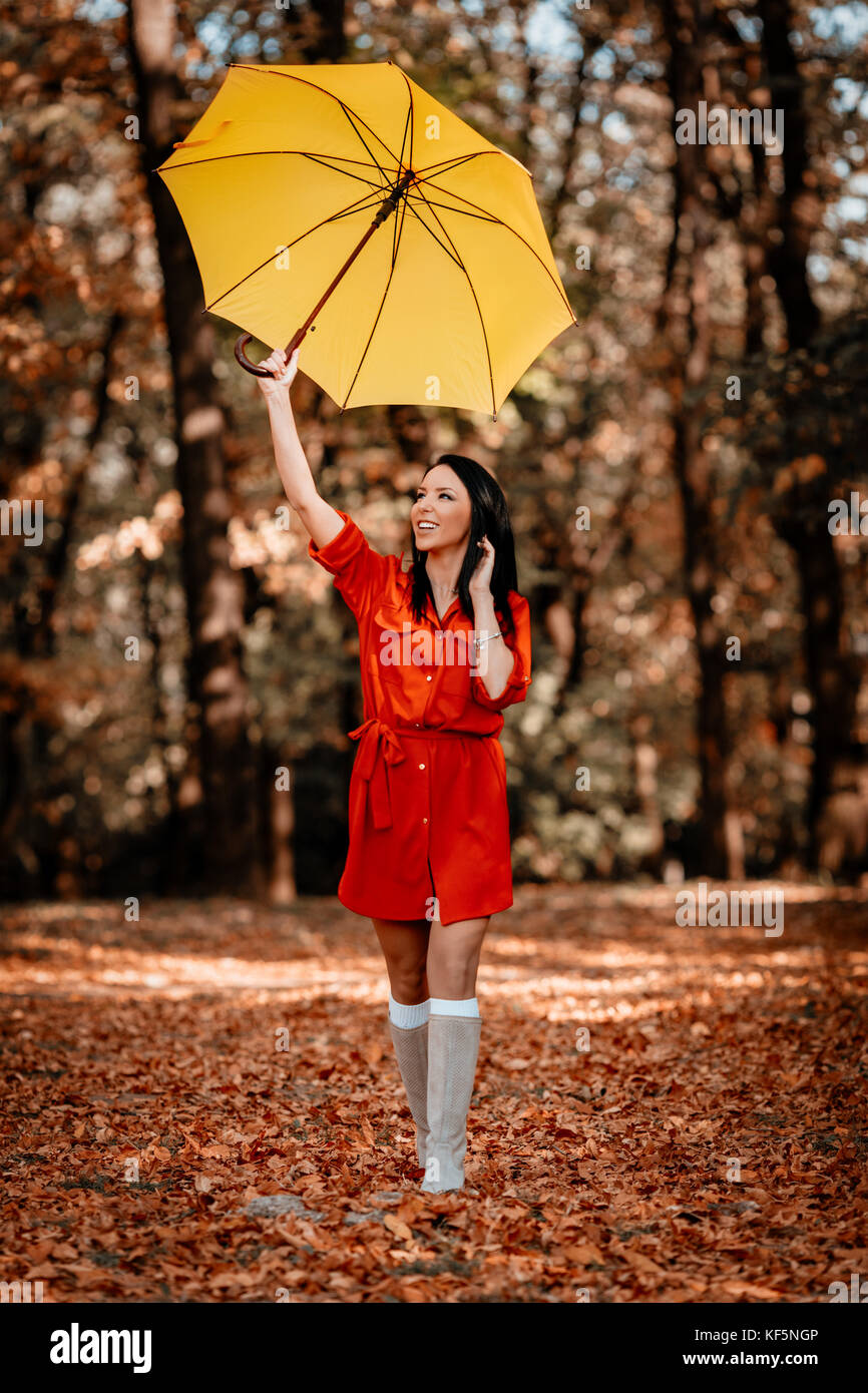 Jeune femme en robe rouge parler sur smartphone balade en automne sunny park, holding parapluie jaune hauteur au-dessus de sa tête. Banque D'Images