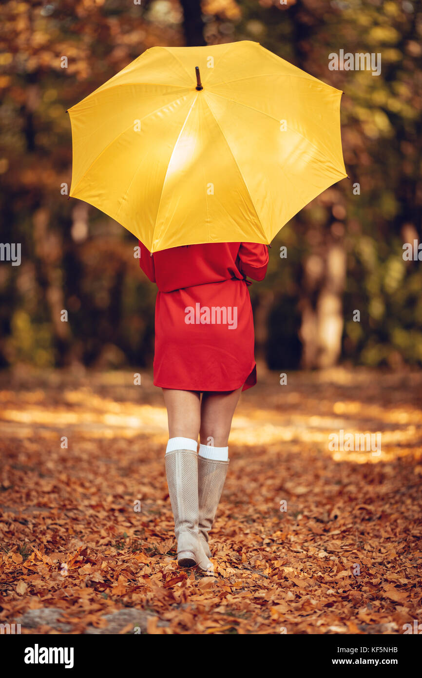 Belle jeune fille en robe rouge aller seul sous parapluie jaune dans le parc en automne doré. vue arrière. Banque D'Images