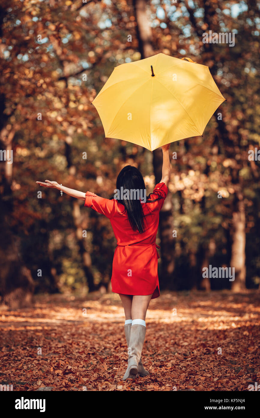 Cheerful young Beautiful woman in red dress aller seul avec parapluie jaune dans le parc en automne doré. vue arrière. Banque D'Images