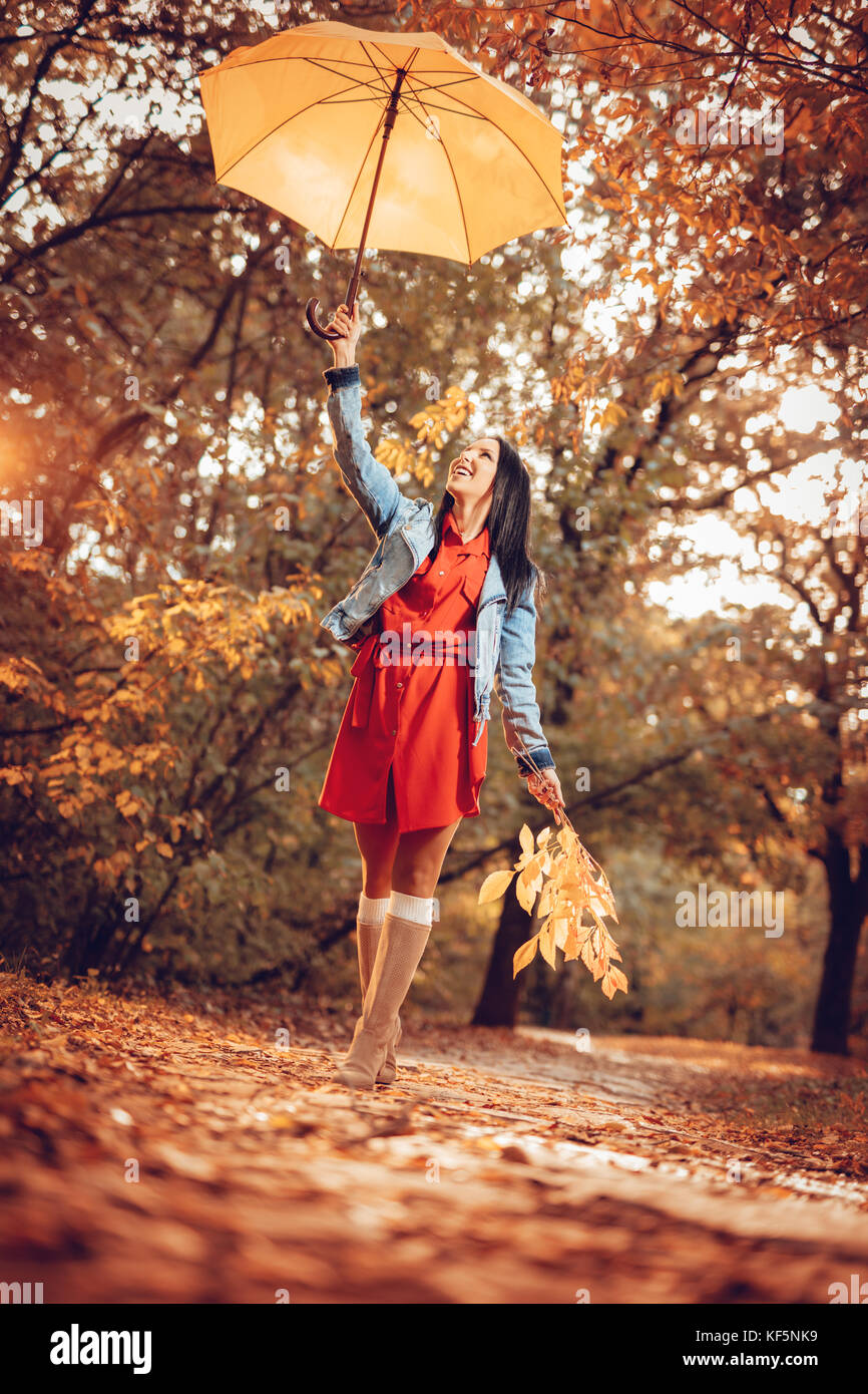 Happy young woman walking in autumn sunny park holding umbrella, hauteur au-dessus de sa tête et une succursale d'une des feuilles tombées dans la main gauche. Banque D'Images