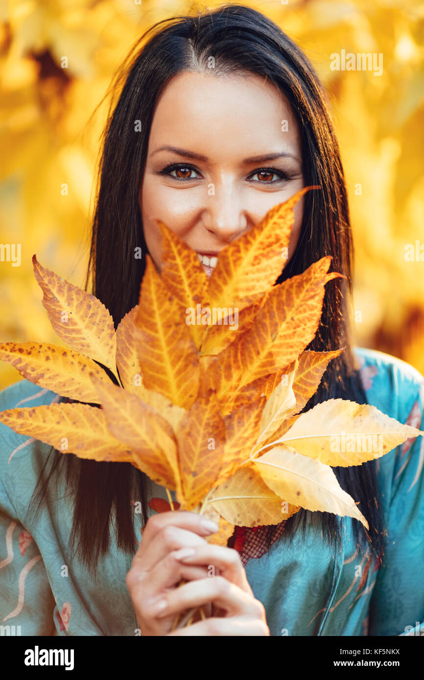 Portrait of beautiful happy woman holding les feuilles d'automne sur son visage dans le parc et souriant. looking at camera. Banque D'Images