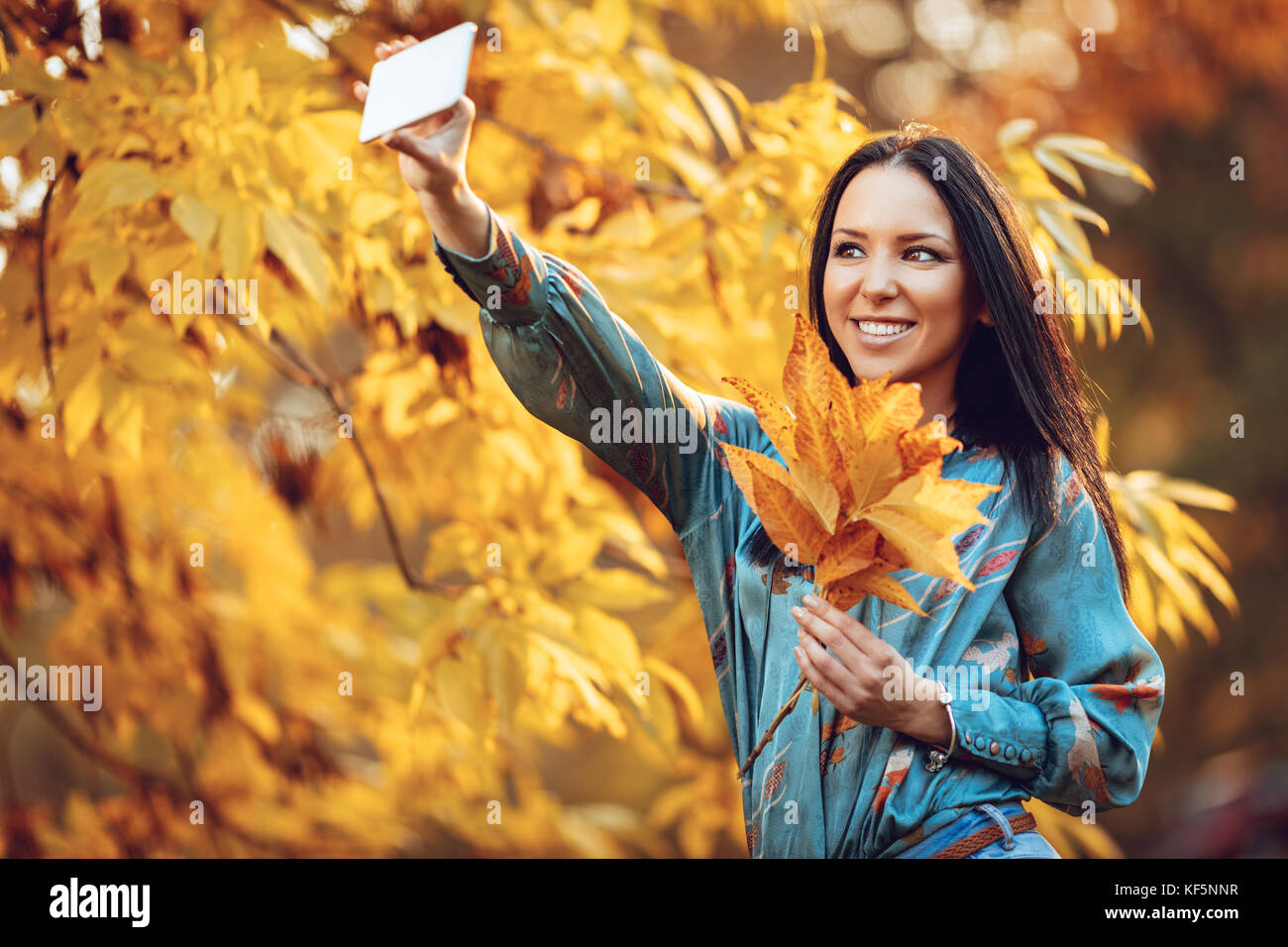 Cute girl moderne prenant un parc en selfies en automne avec des feuilles sèches dans sa main. Banque D'Images