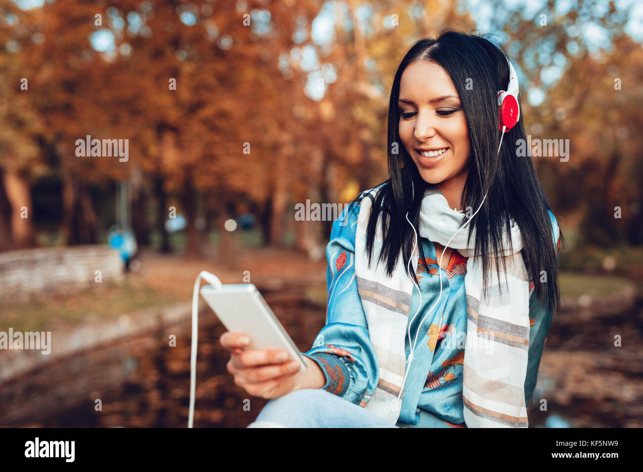 Jeune femme regarder des vidéos et écouter de la musique sur un smartphone avec le casque sur ses oreilles et assis dans un parc sur une magnifique journée d'automne. Banque D'Images