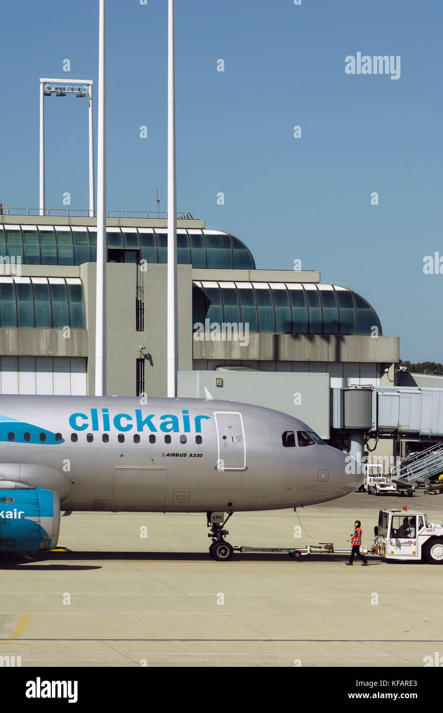 Avant du fuselage d'un Airbus A320-200 clickair, un remorqueur avec un attelage, un travailleur de l'aéroport orange portant un tabard et ear-humains avec une passerelle et te Banque D'Images