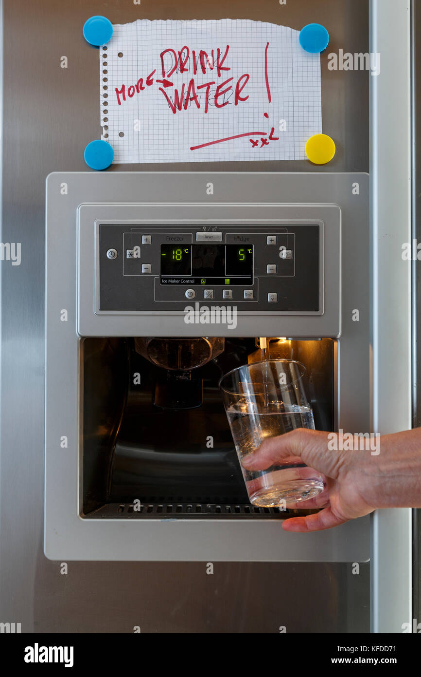 Une main tenant un verre sous le distributeur d'eau d'un frigo américain  avec un 'drink plus d'eau" part écrit sur papier collé sur le frigo avec  des aimants de réfrigérateur Photo Stock -