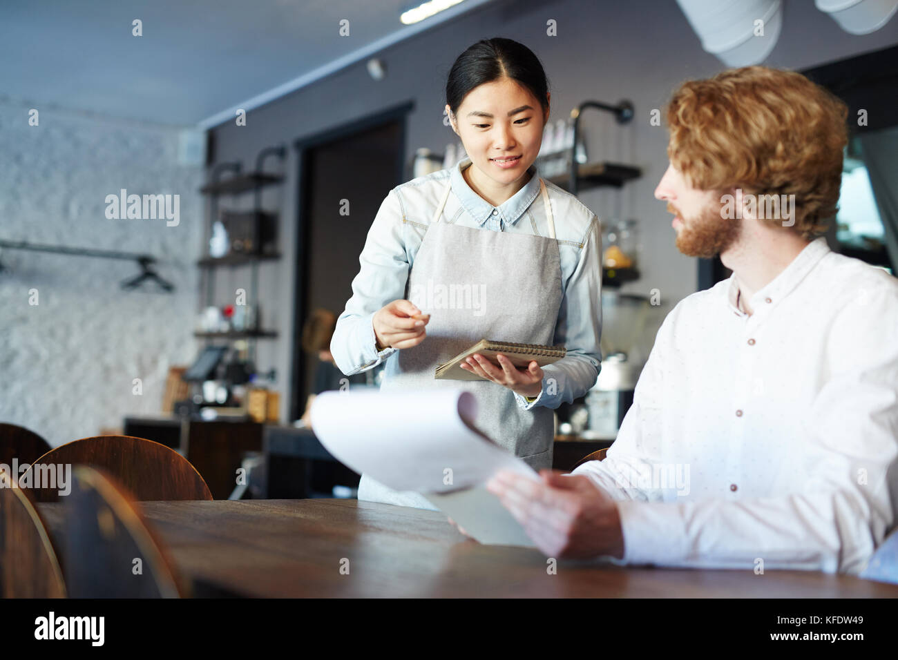 Jeune serveuse asiatique de conseiller l'un des clients de choisir nouveau snack à partir d'un menu dans un restaurant Banque D'Images