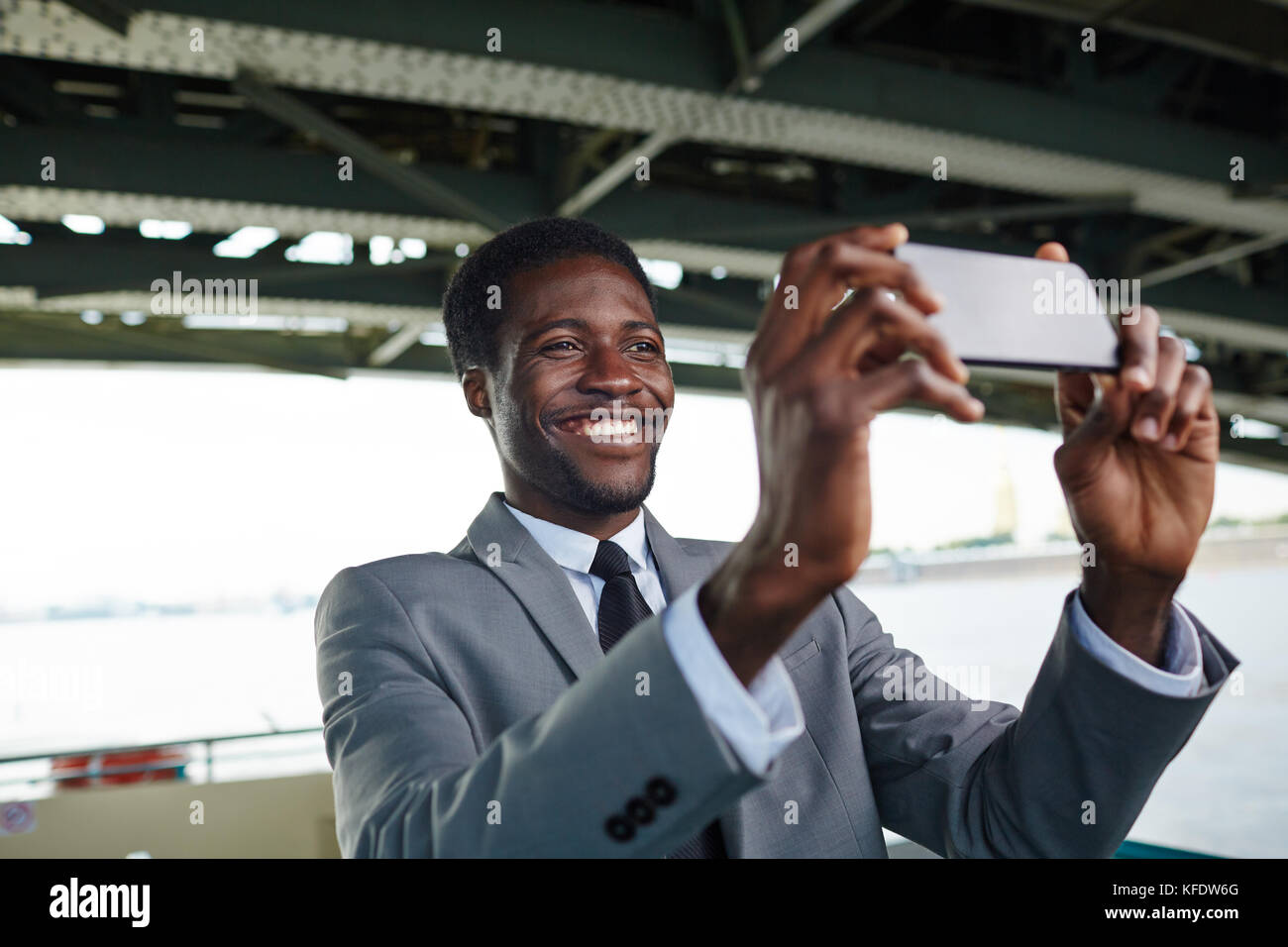 Cheerful african american entrepreneur dans le costume classique en tenant sur smartphone selfies en se tenant sur le pont supérieur du navire, l'arrière-plan flou Banque D'Images