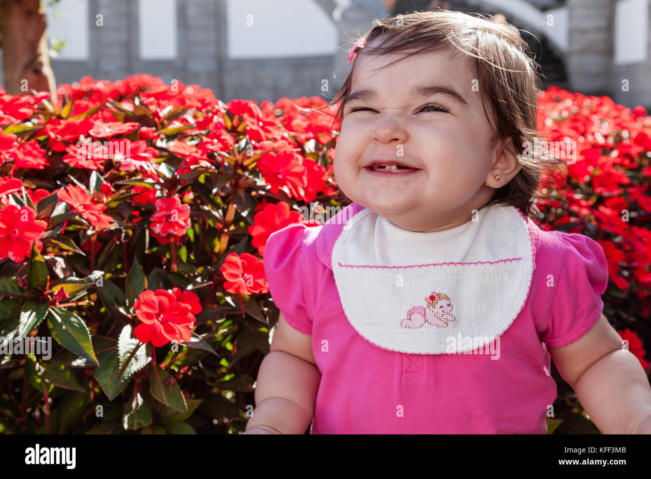 Mignon, souriant, heureux, joli bébé joufflu avec un grand sourire, rire et montrant deux dents dans un jardin de fleurs rouges. Quatorze mois / baby Banque D'Images