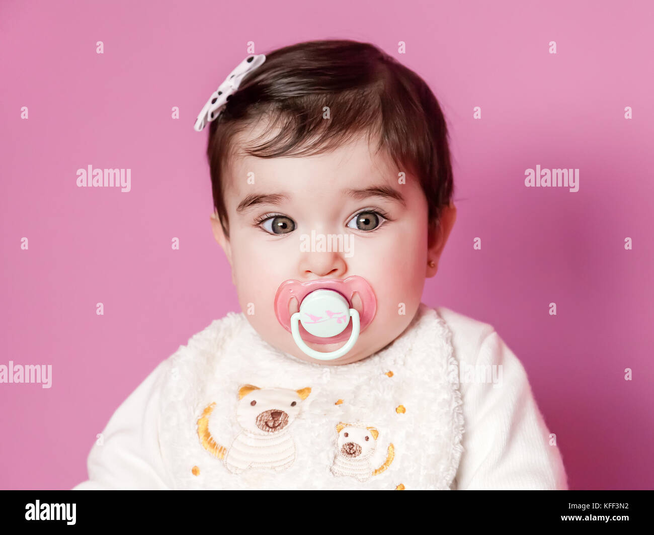 Mignon, joli et happy baby girl portrait avec une sucette rose, mannequin ou sucette, portant un dossard et un arc dans les cheveux. neuf mois / mannequin bébé fille Banque D'Images