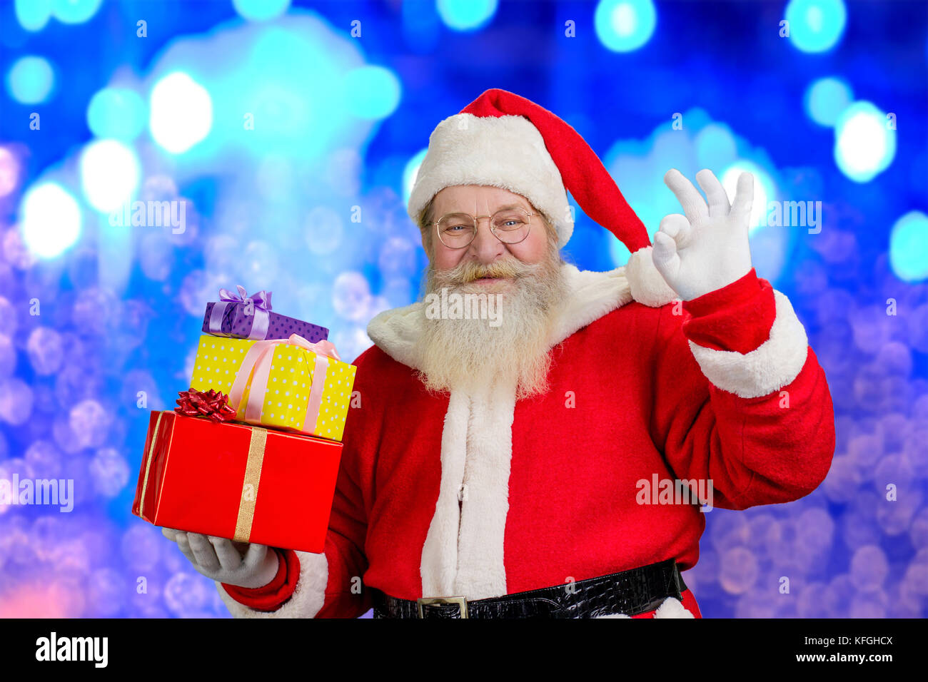 Le père Noël avec les cadeaux de noël santa claus holding réaliste. des boîtes à cadeaux et montrant sign ok, bleu fond brillant. Banque D'Images