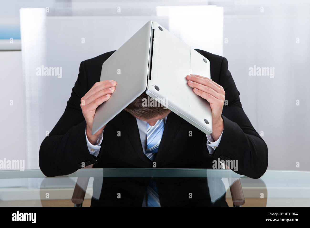 Déprimé businessman sitting at a table réfléchissant cachait sous son ordinateur portable comme il le maintient ouvert sur sa tête s'inclina Banque D'Images