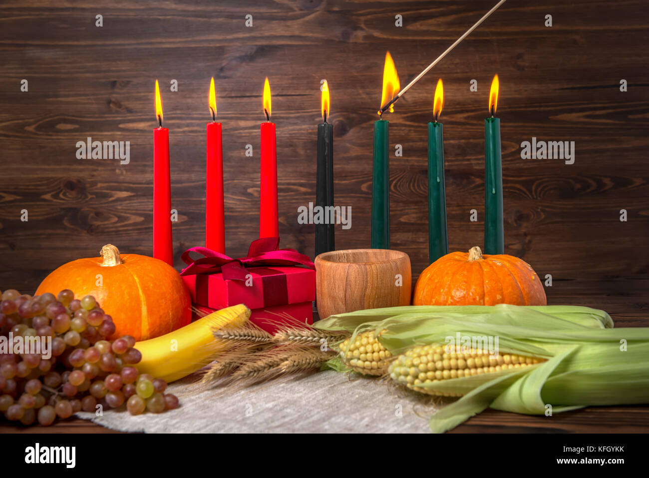 L'allumage des bougies traditionnelles Kwanzaa, festival concept avec boîte-cadeau, citrouilles, épis de blé, raisins, maïs, bananes, fruits et bol en bois sur bac Banque D'Images