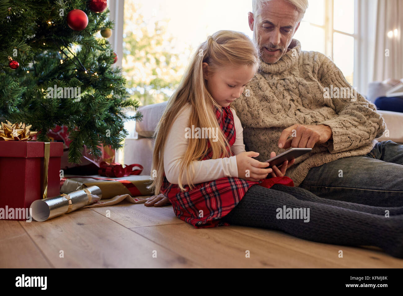 Petite fille avec son grand-père assis par arbre de Noël à la maison et à l'aide de tablette numérique. Assis tous les deux sur le plancher dans la salle de séjour et looking at tablet pc Banque D'Images