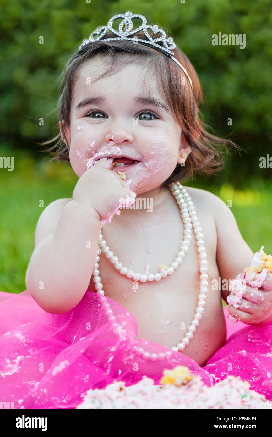 Smiling happy baby girl premier anniversaire anniversaire partie. lécher les doigts avec du rose sale face gâteau. princess tiara costume Banque D'Images