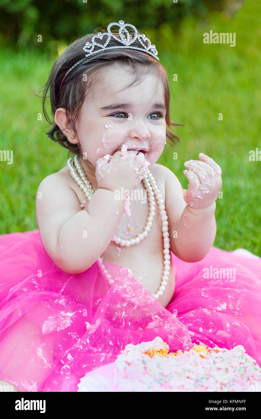 Smiling happy baby girl premier anniversaire anniversaire partie. lécher la main avec sale face de gâteau rose costume princesse tiara. Banque D'Images