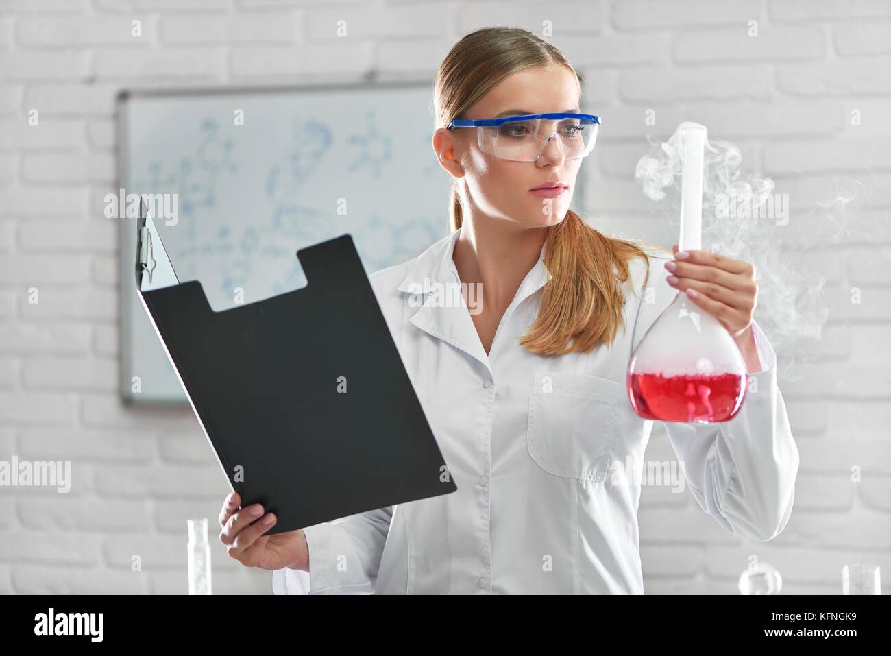 Tiré d'une belle femme portant des lunettes de scientifique travaillant dans son laboratoire holding clipboard et une fiole avec de l'échantillon chimique fumeurs reacti Banque D'Images