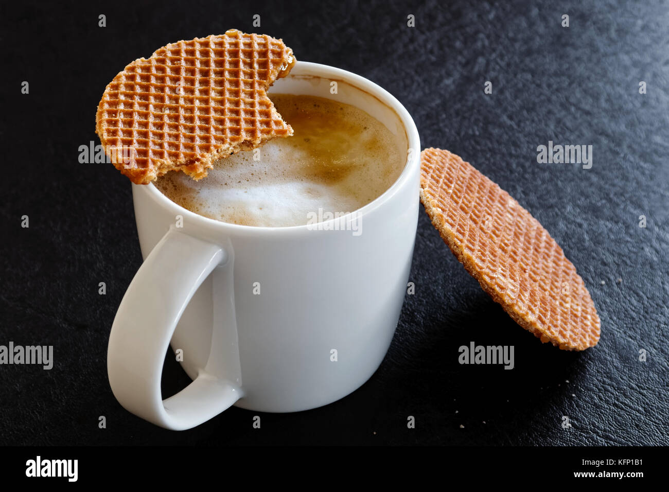 Mug blanc laiteux avec délicieuse partiellement ivre et partiellement mangés un biscuit gaufre sur le dessus. fond noir. Banque D'Images