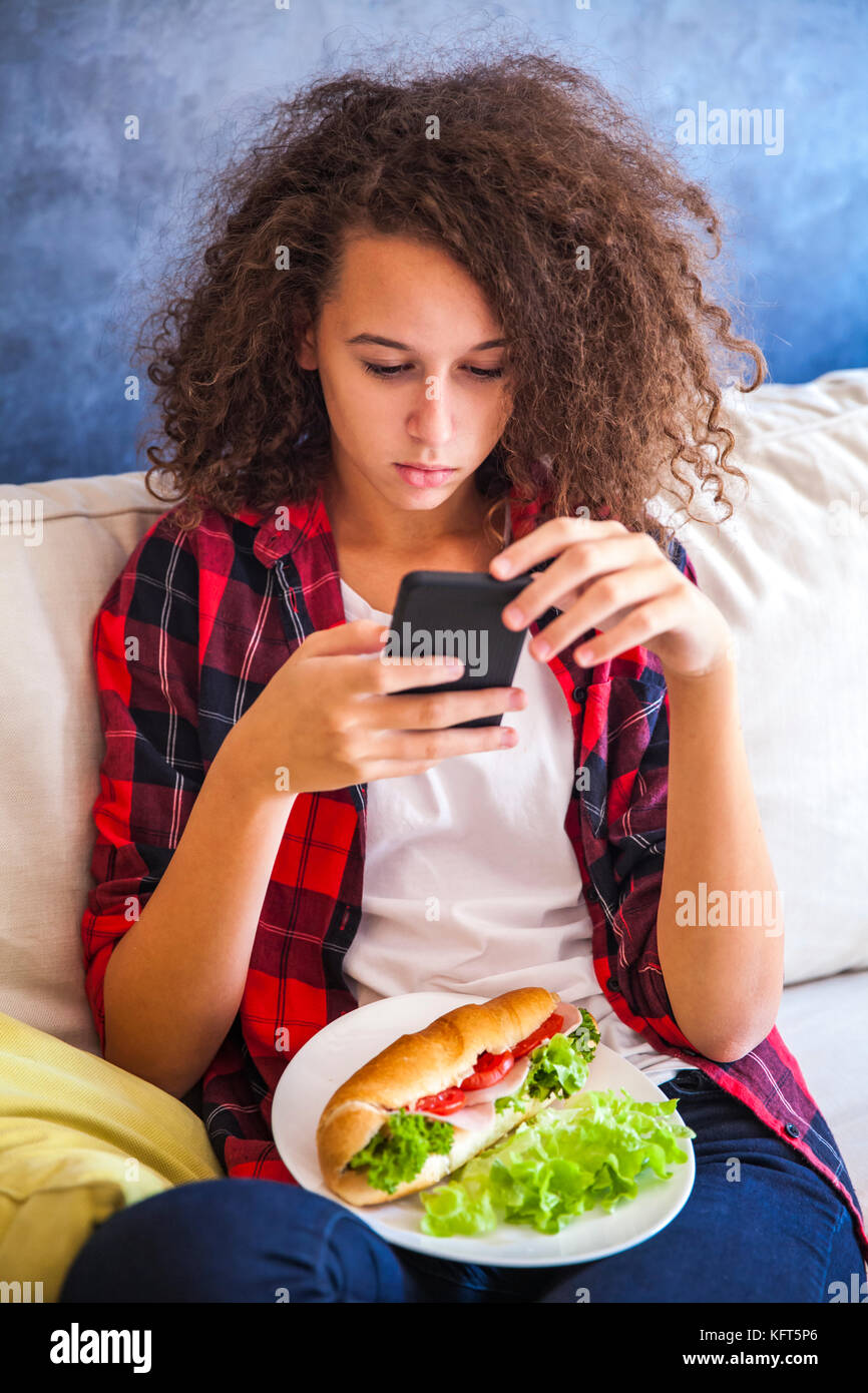 Cheveux bouclés teen girl à l'aide téléphone mibile et eating sandwich sur canapé Banque D'Images