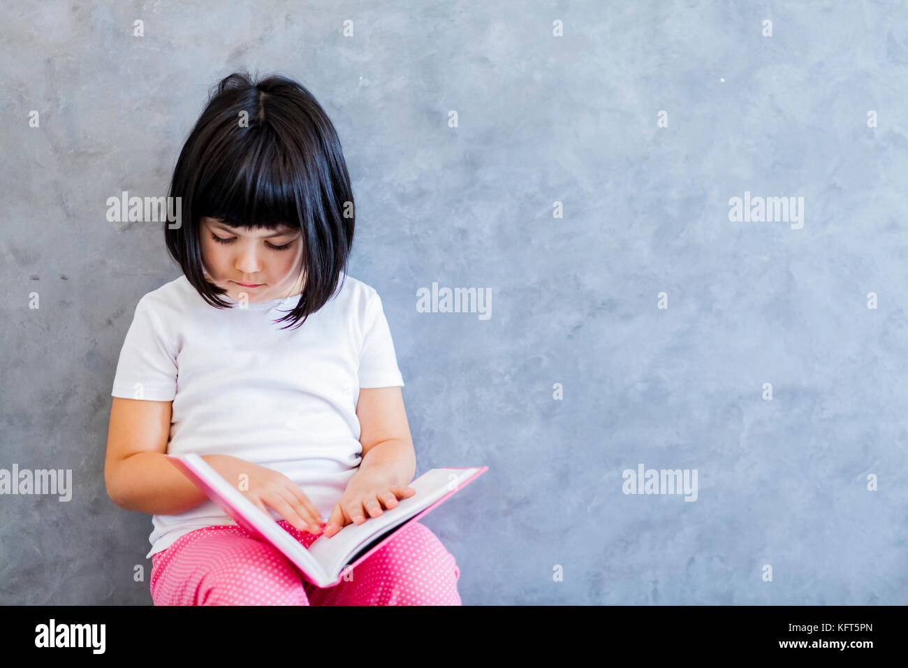 Portrait of cute little girl cheveux noir livre de lecture par le mur Banque D'Images