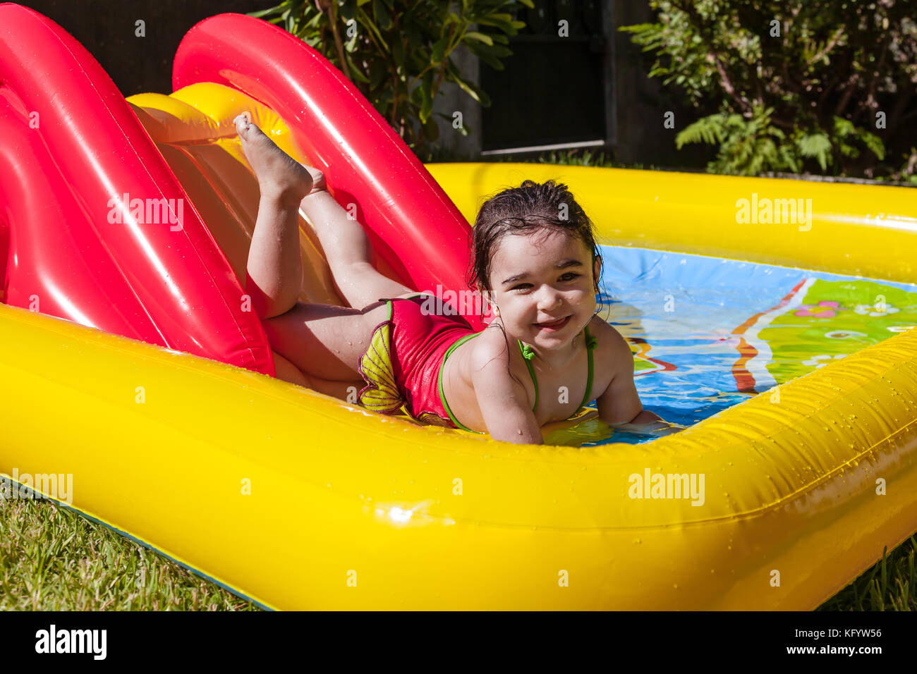 Heureux, souriant tout-petit, jouant et éclaboussant dans le curseur de la piscine gonflable colorée de jardin d'arrière-cour dans la journée chaude d'été. Treize mois Banque D'Images