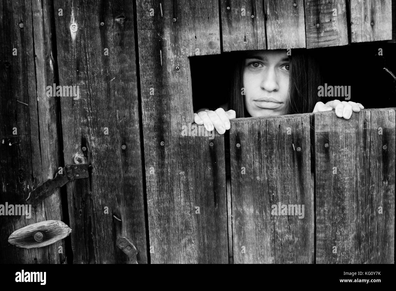 Jeune femme à la recherche à travers la fente d'un hangar en bois. photo en noir et blanc. Banque D'Images