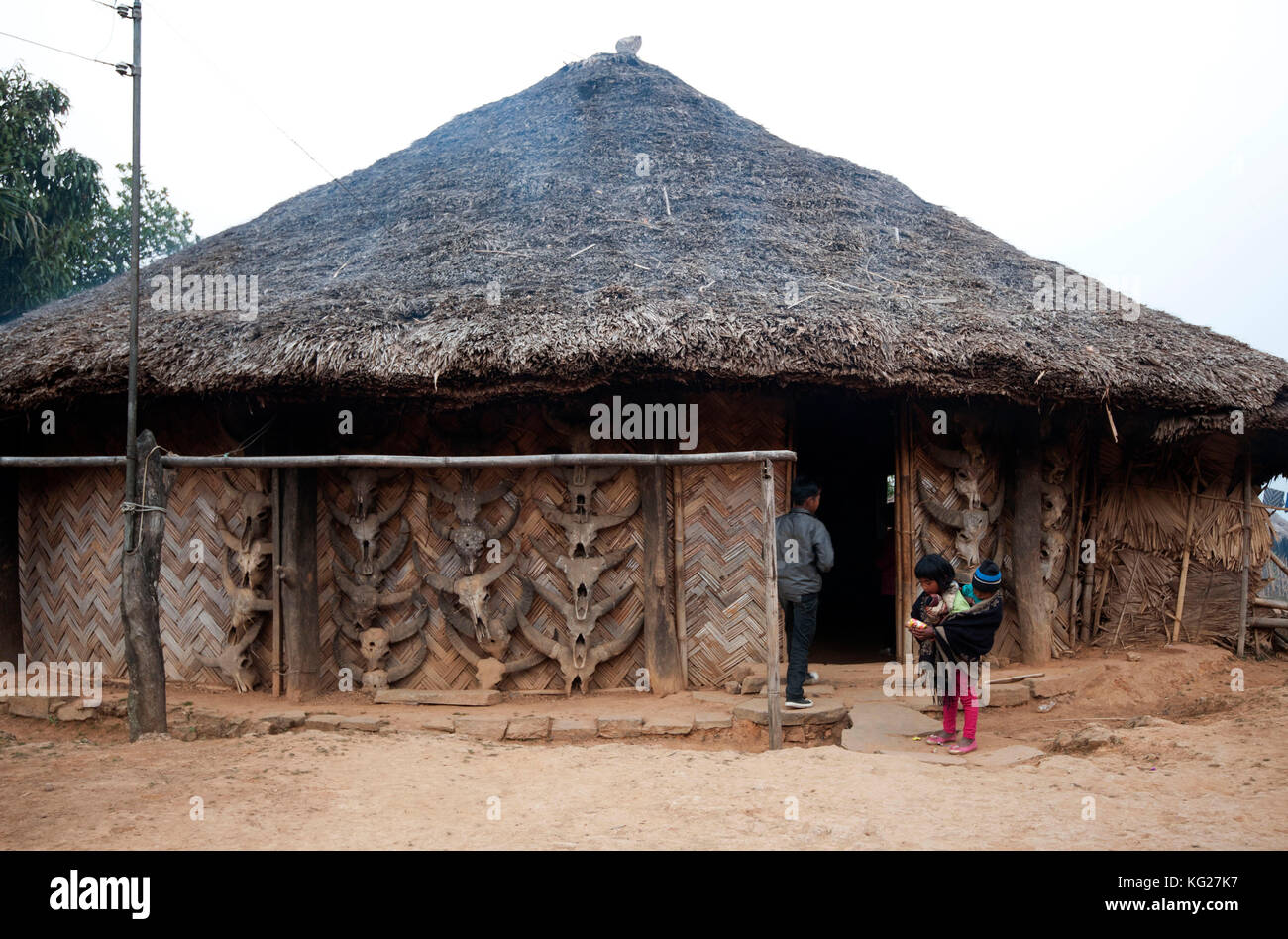 Jeune fille portant des frères et sœurs en papoose en tissu à l'extérieur d'une maison de chaume de palmier construite traditionnellement, avec des crânes de Mithun, Nagaland, Inde, Asie Banque D'Images