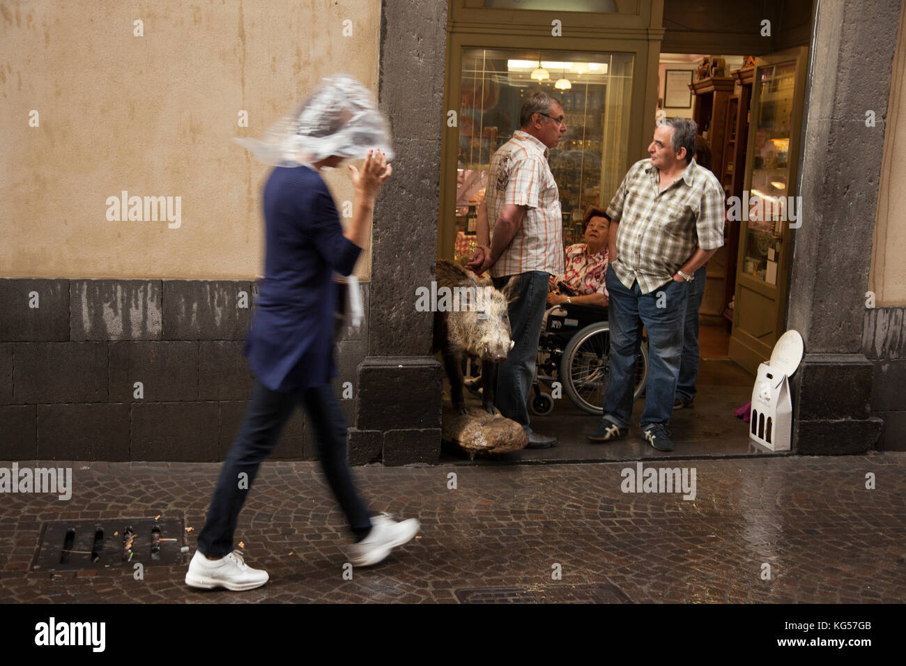 Une femme porte sur sa tête en plastique comme elle passe devant une boucherie dans la pluie dans la ville ombrienne de Orvieto, Italie. Banque D'Images