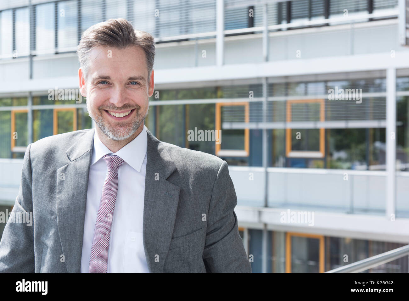 Businessman sur balcon en face de l'immeuble de bureaux, l'air dans l'appareil photo en souriant, portrait Banque D'Images