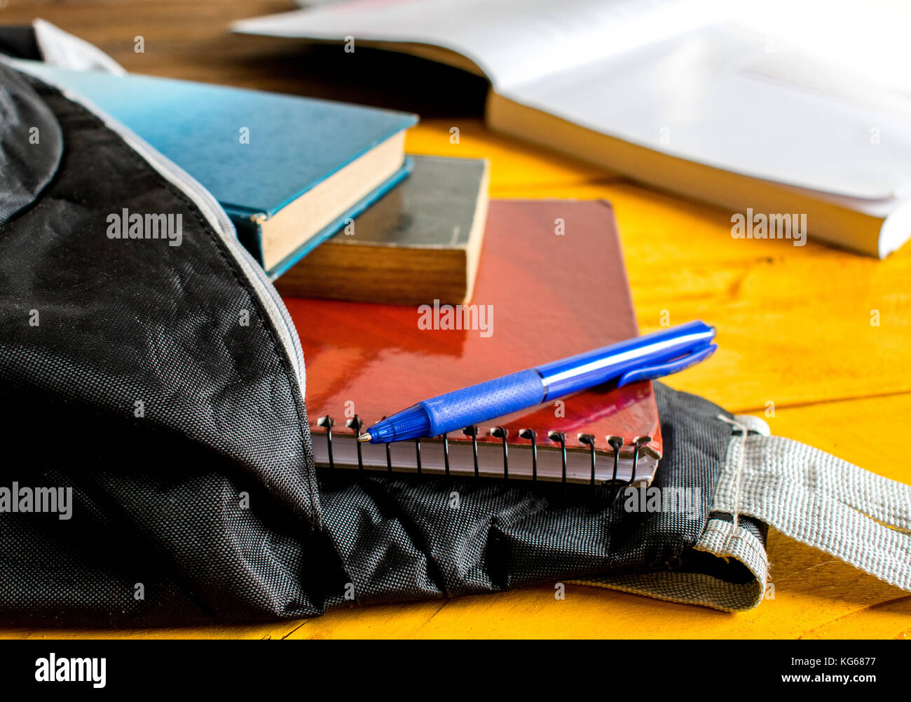 Sac à dos noir école ouverte, ou un sac, avec trois livres et cahiers et stylo bleu légèrement déversant hors de lui, en haut d'une surface de table en bois jaune Banque D'Images