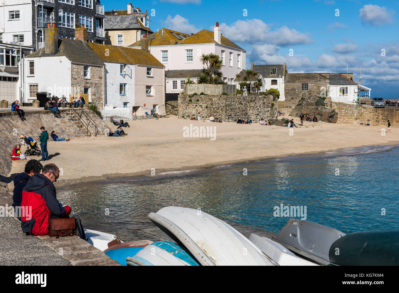 Saint Ives, Angleterre - 27 avril 2017 : touristes assis au soleil sur le quai du port d'Ives avec bateaux, eau claire, plage et maisons. Banque D'Images