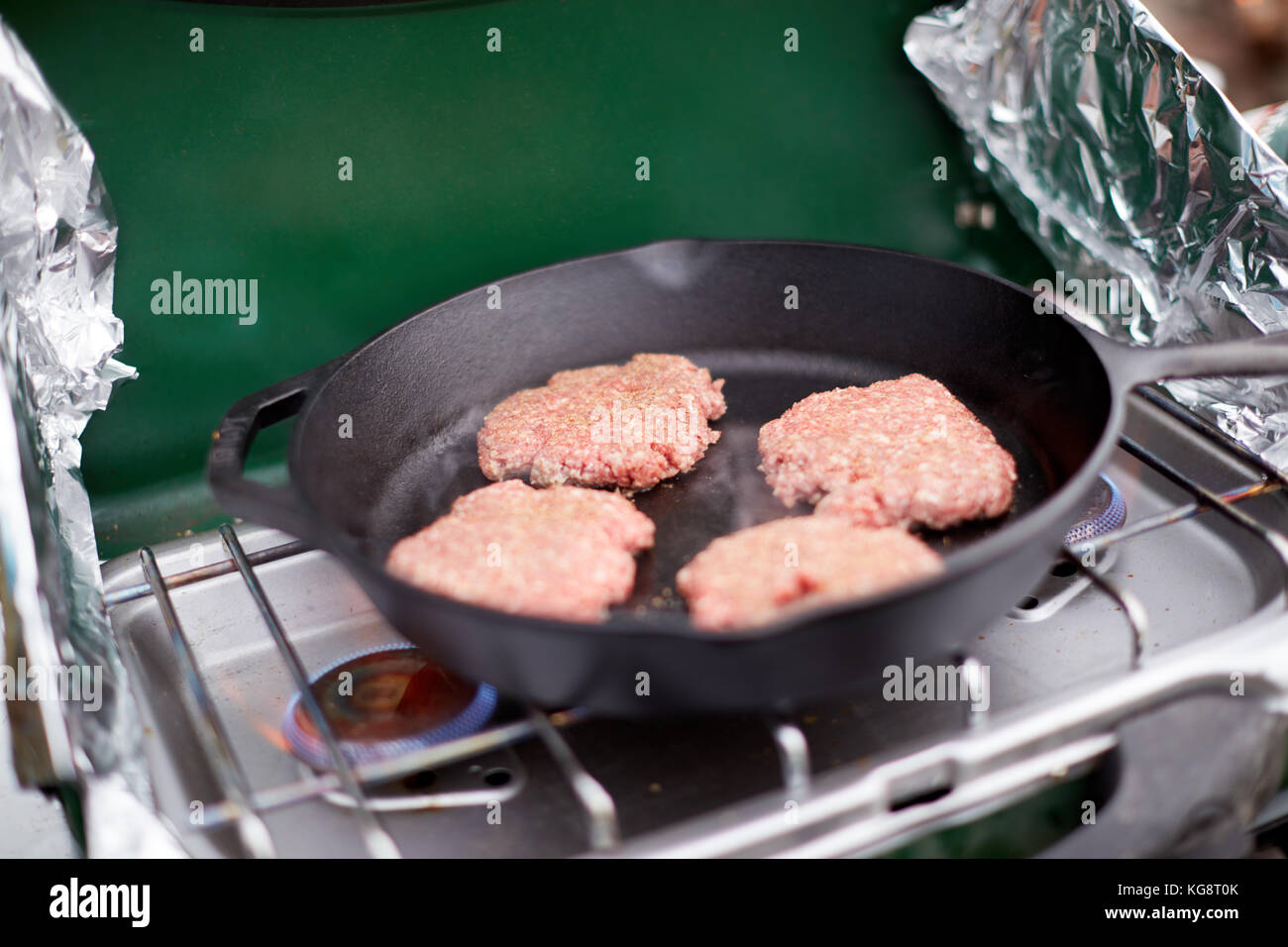 Des galettes de boeuf haché cru dans un poêlon sur un réchaud à gaz de camping d'un papier aluminium pendant la cuisson à l'extérieur des gardes du vent Banque D'Images