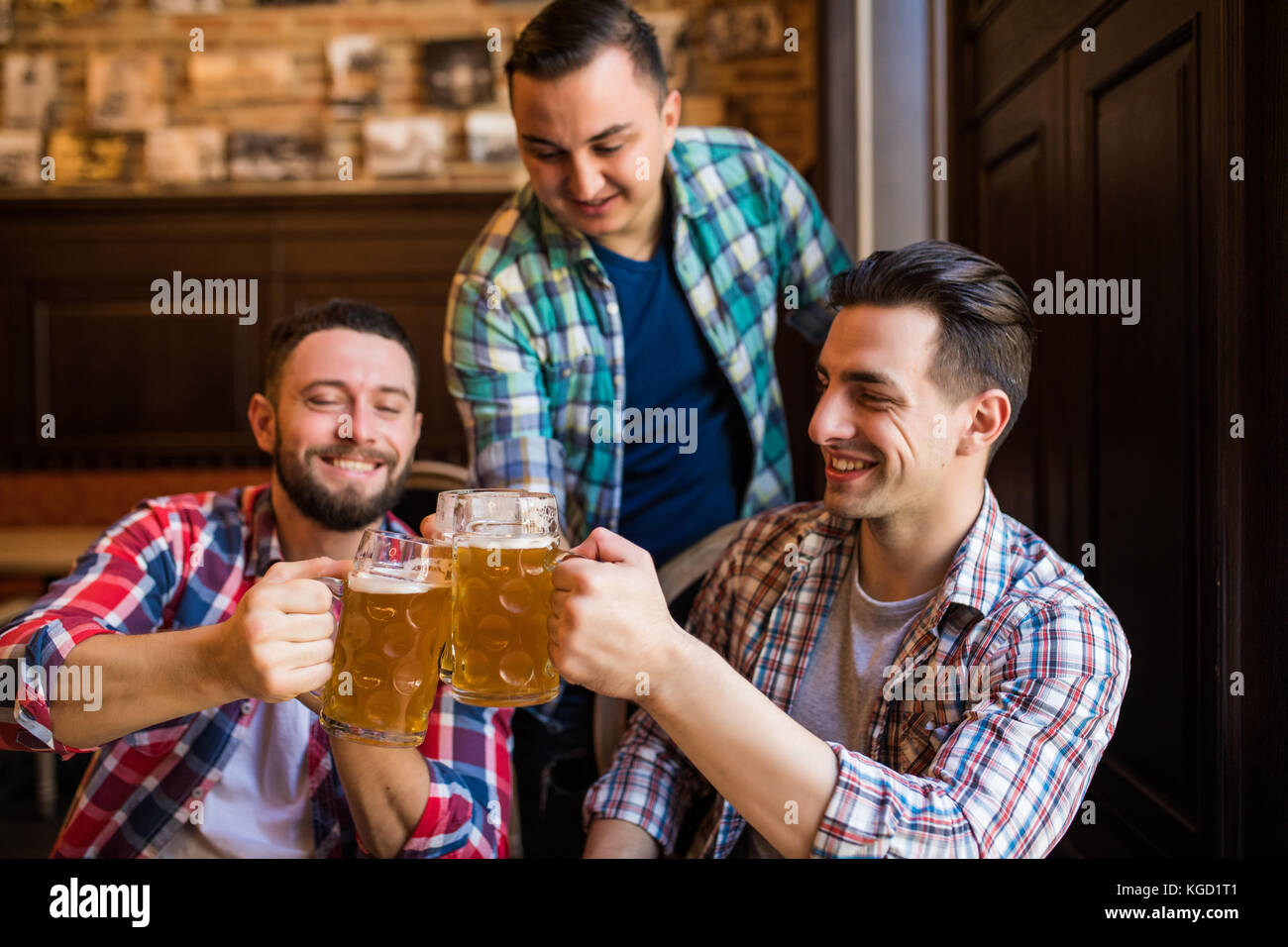 Trois jeunes hommes en vêtements décontractés sont souriants qui retentit et verres de bière ensemble tout en restant assis dans un pub. de la bière. Banque D'Images