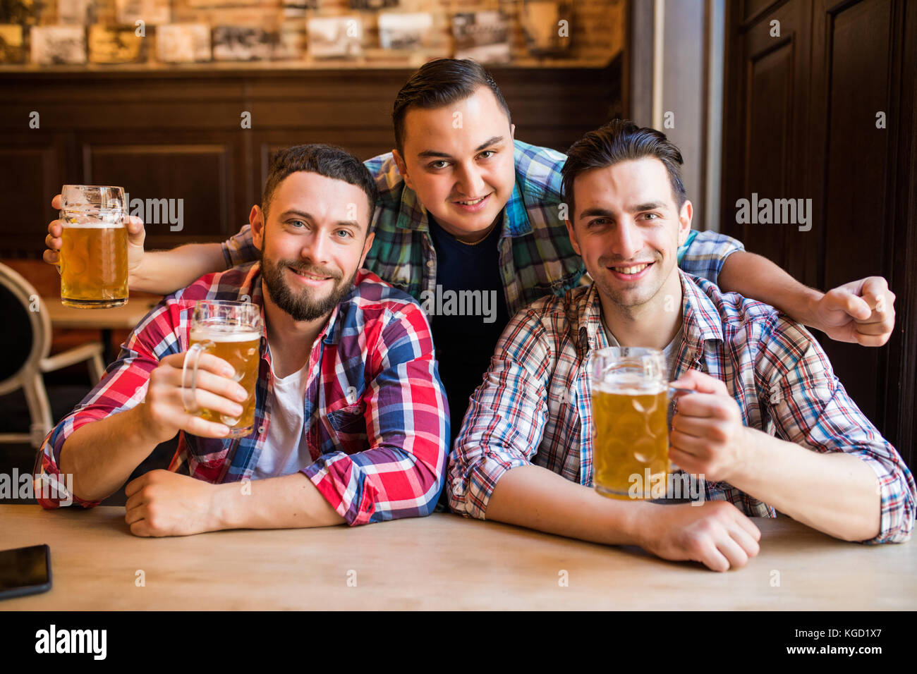 Trois jeunes hommes en vêtements décontractés sont souriants qui retentit et verres de bière ensemble tout en restant assis dans un pub. de la bière. Banque D'Images