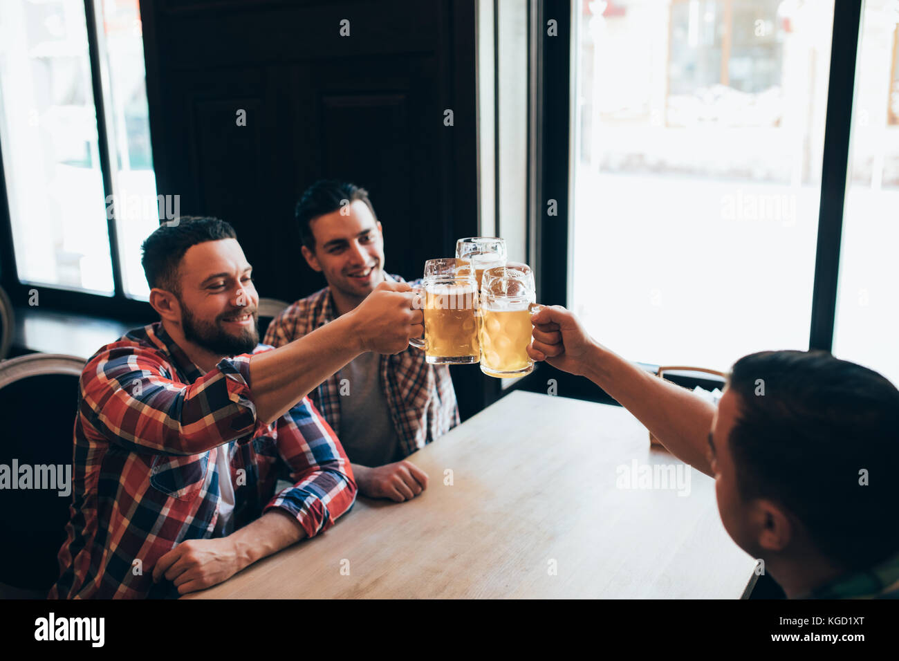 Trois jeunes hommes en vêtements décontractés sont souriants qui retentit et verres de bière ensemble tout en restant assis dans un pub Banque D'Images
