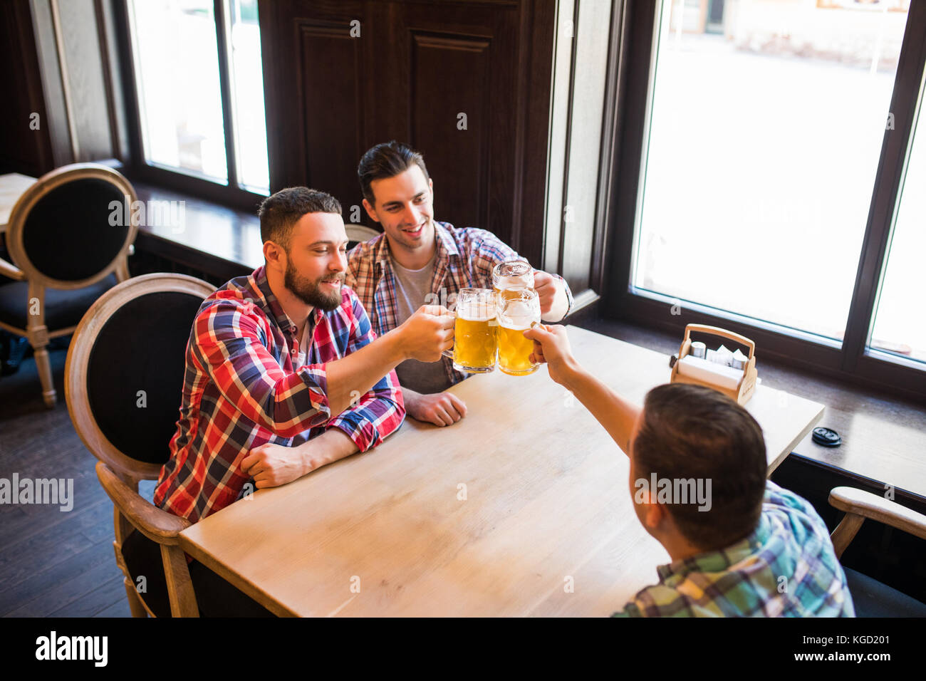 Trois jeunes hommes en vêtements décontractés sont souriants qui retentit et verres de bière ensemble tout en restant assis dans un pub Banque D'Images
