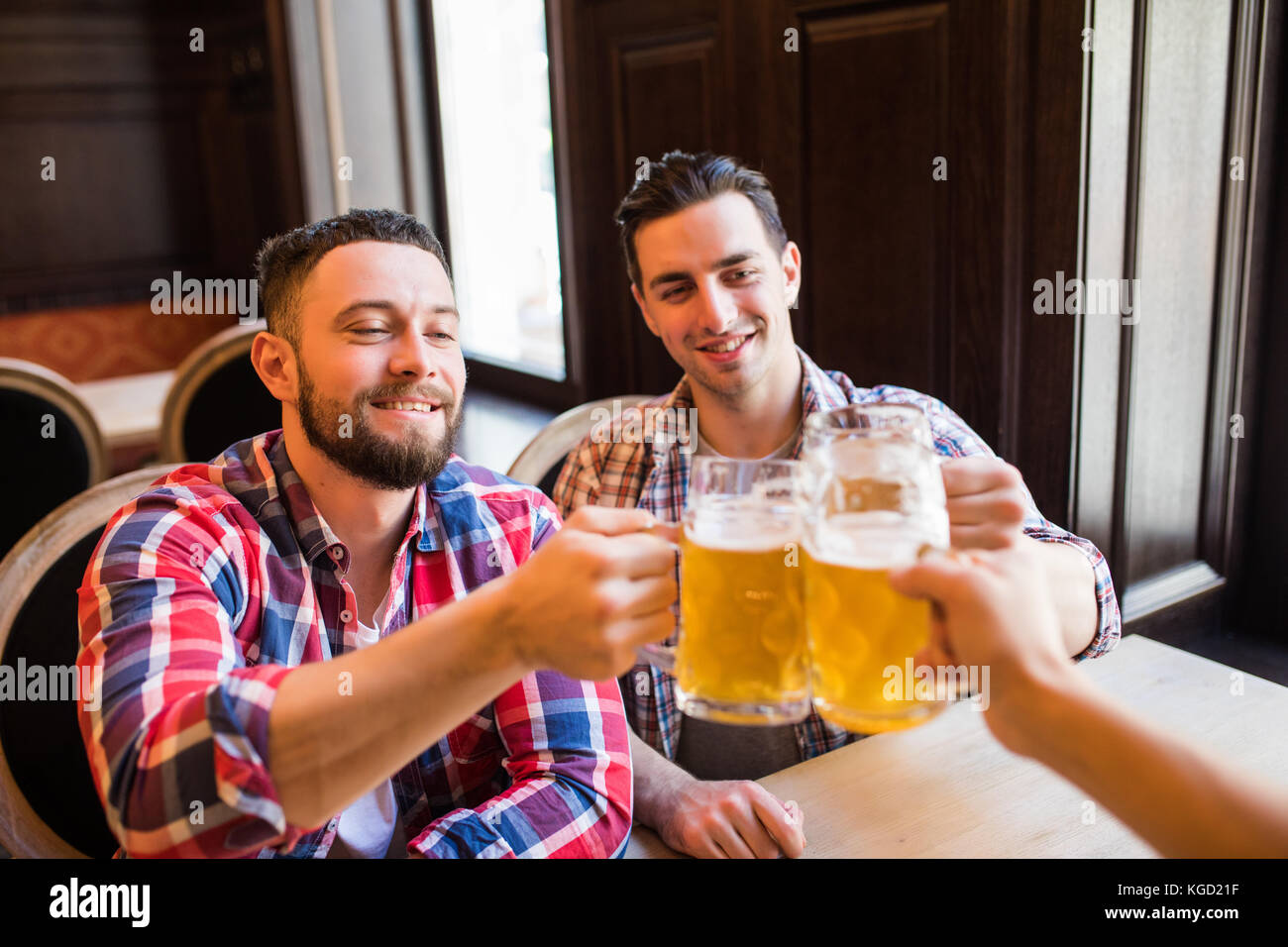 Trois jeunes hommes en vêtements décontractés sont souriants qui retentit et verres de bière ensemble tout en restant assis dans un pub Banque D'Images