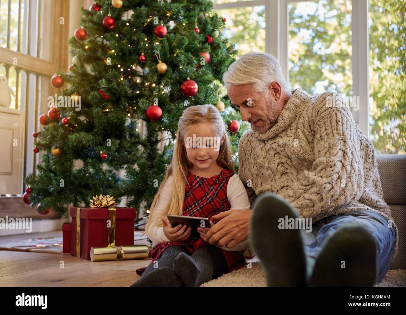 Senior man with granddaughter using digital tablet, assis tous les deux près de l'arbre de Noël. Banque D'Images