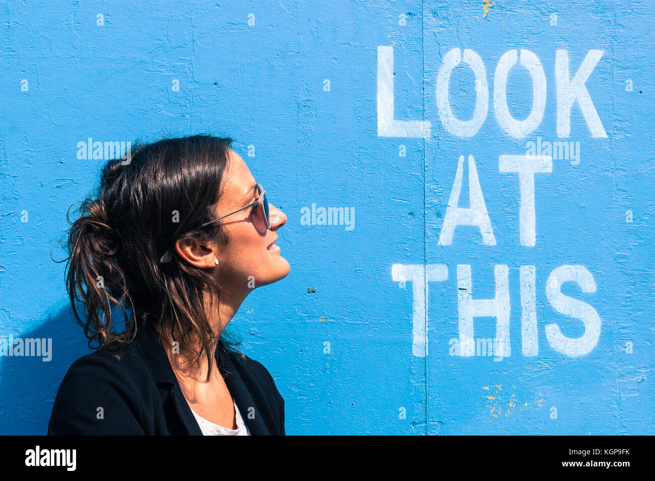 Portrait d'un modèle portant des lunettes de soleil qui pose à côté d'un mur bleu avec les mots 'regardez ce' écrit dessus Banque D'Images