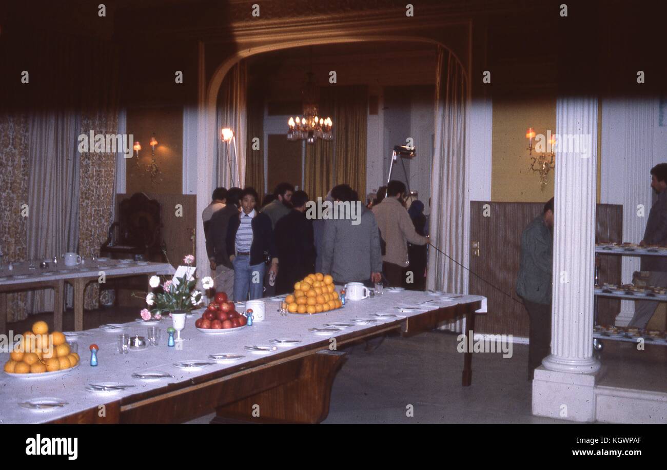 Scène d'une longue table de banquet avec couverts et grandes assiettes de fruits, dans un hôtel en Iran, mars 1983. En arrière-plan, un groupe de reporters et de cameramen filmant quelque chose d'invisible dans l'autre pièce. Dommages importants sur la photo originale. () Banque D'Images