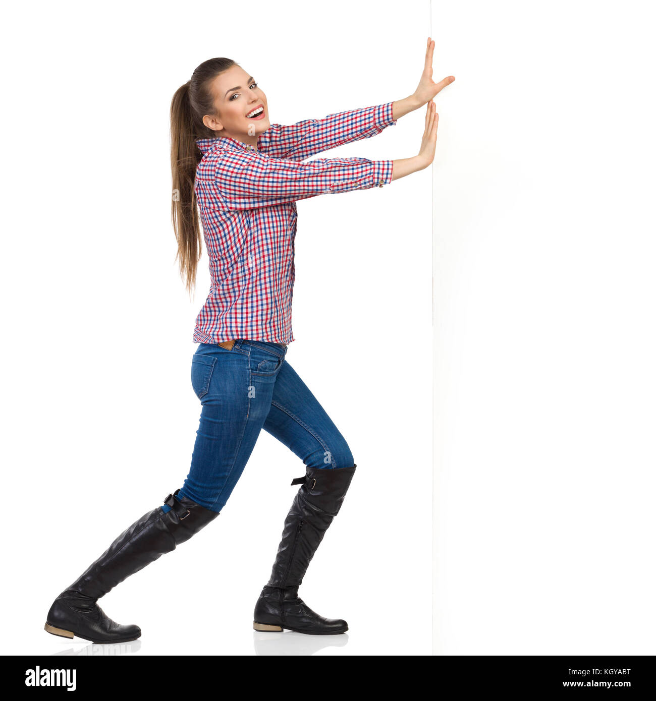 Jeune femme en jeans, bottes noires et chemise de bûcheron poussant un mur blanc et à la recherche à l'appareil photo. Vue de côté, pleine longueur isolée studio shot Banque D'Images
