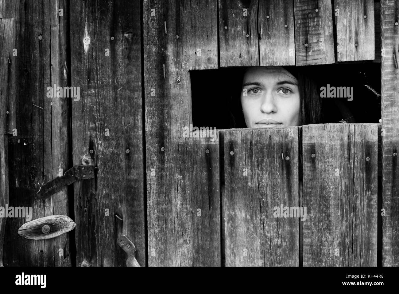 Une jeune femme enfermée dans une cabane en bois, peeking through une petite fenêtre. noir et blanc photo. Banque D'Images